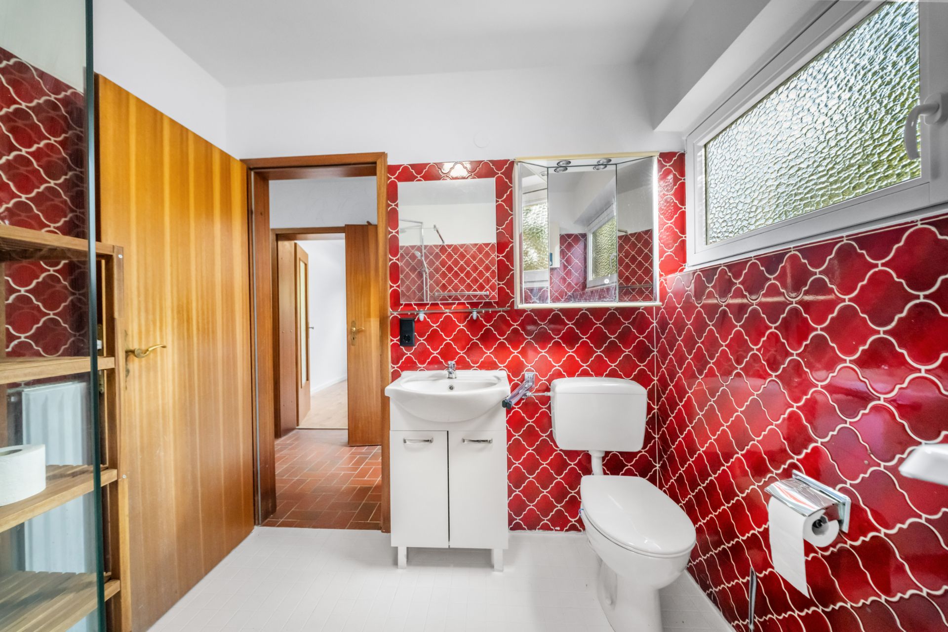 Badezimmer mit roten Fliesen, weißem Waschbecken, Toilette und Fenster.