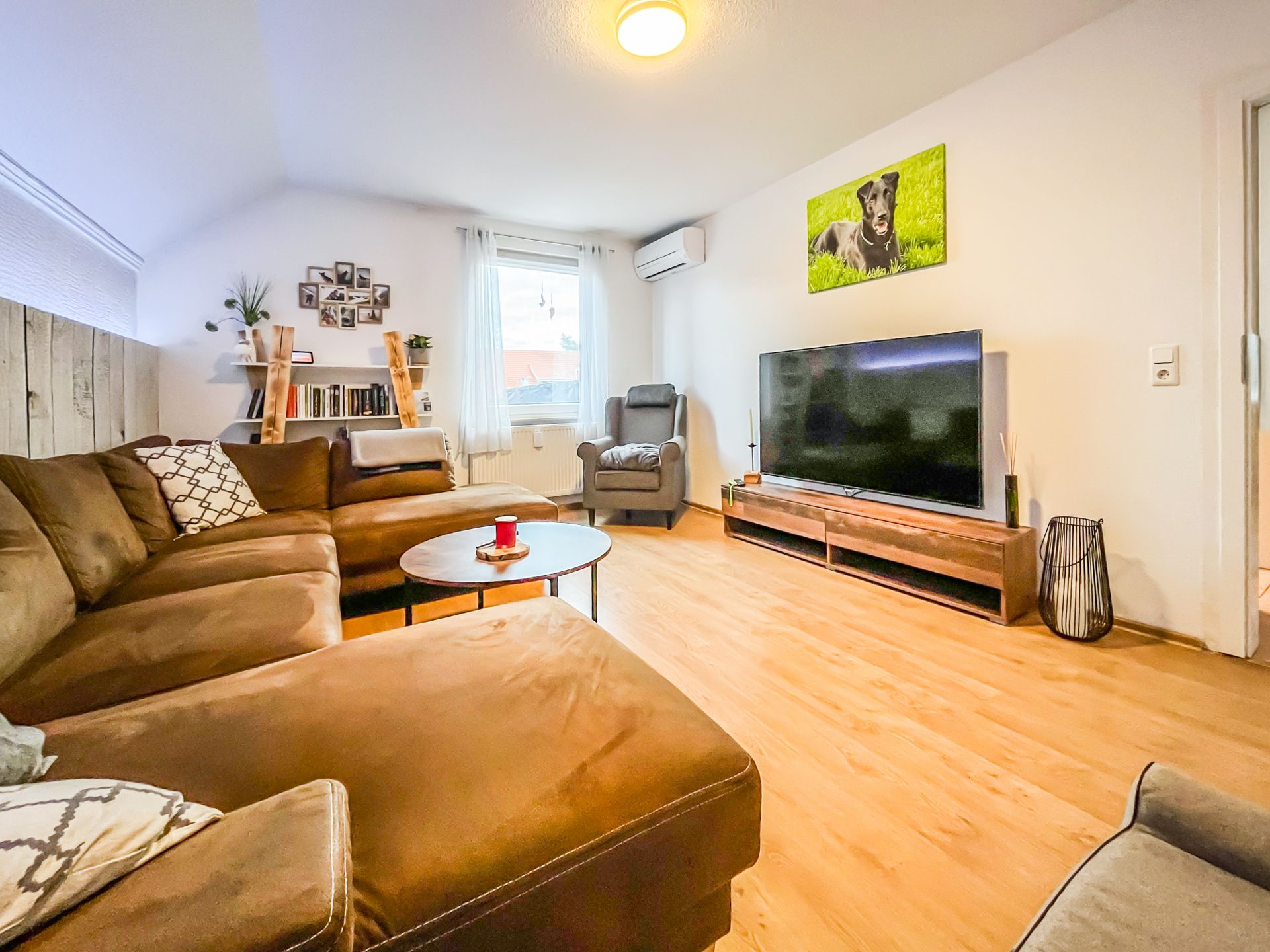 Wohnzimmer mit braunem Ecksofa, Couchtisch, Fernseher und Regal, helles Fenster.