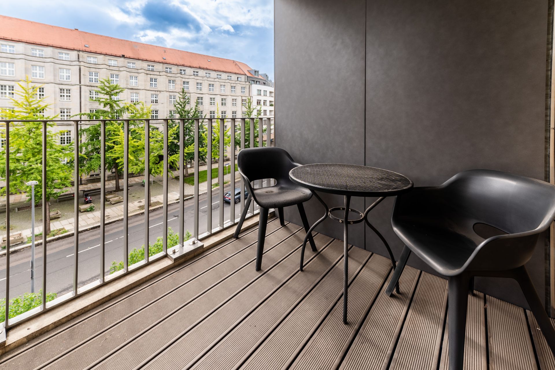 Balkon mit zwei schwarzen Stühlen und rundem Tisch, Blick auf Straße und Gebäude.