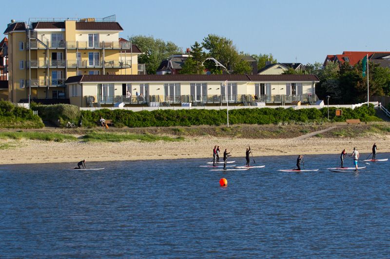 Wohngebäude am Strand mit mehreren Menschen, die auf dem Wasser Stand Up Paddling machen.