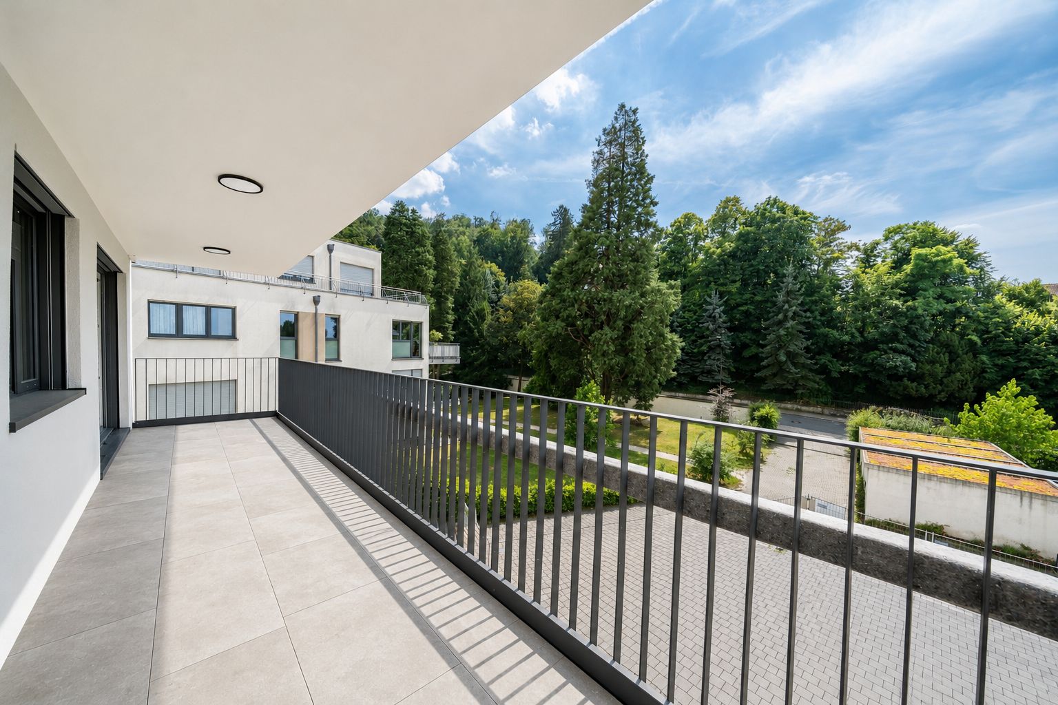 Großer Balkon mit Fliesenboden und Blick auf Bäume und eine ruhige Wohnanlage.