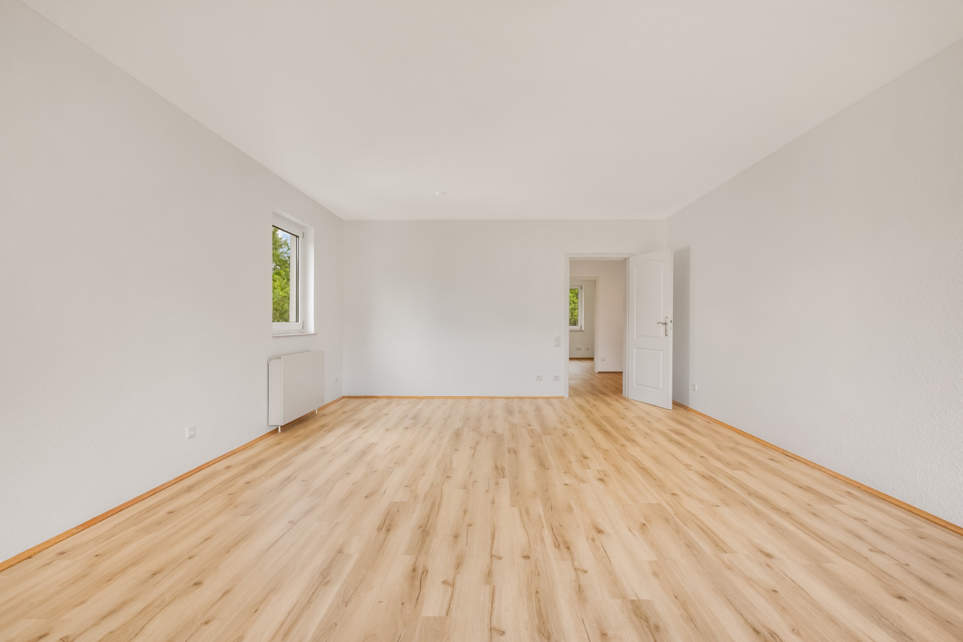Leerer Raum mit hellem Holzboden, weißer Wand, Fenster und geöffneter Tür.