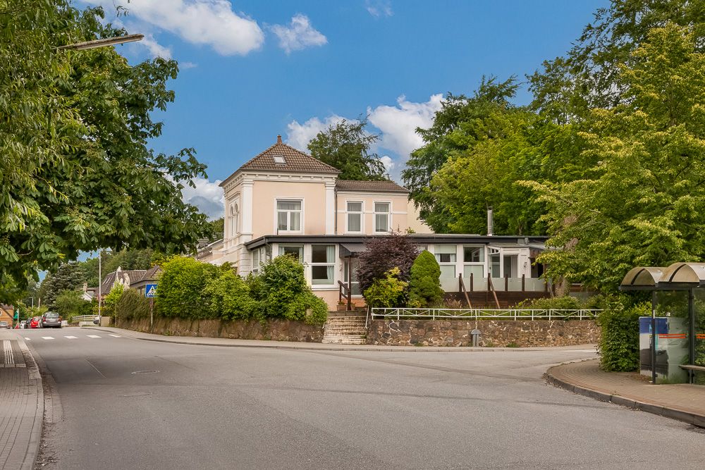 Wohnhaus mit weißer Fassade, umgeben von Bäumen, an einer Straßenecke.