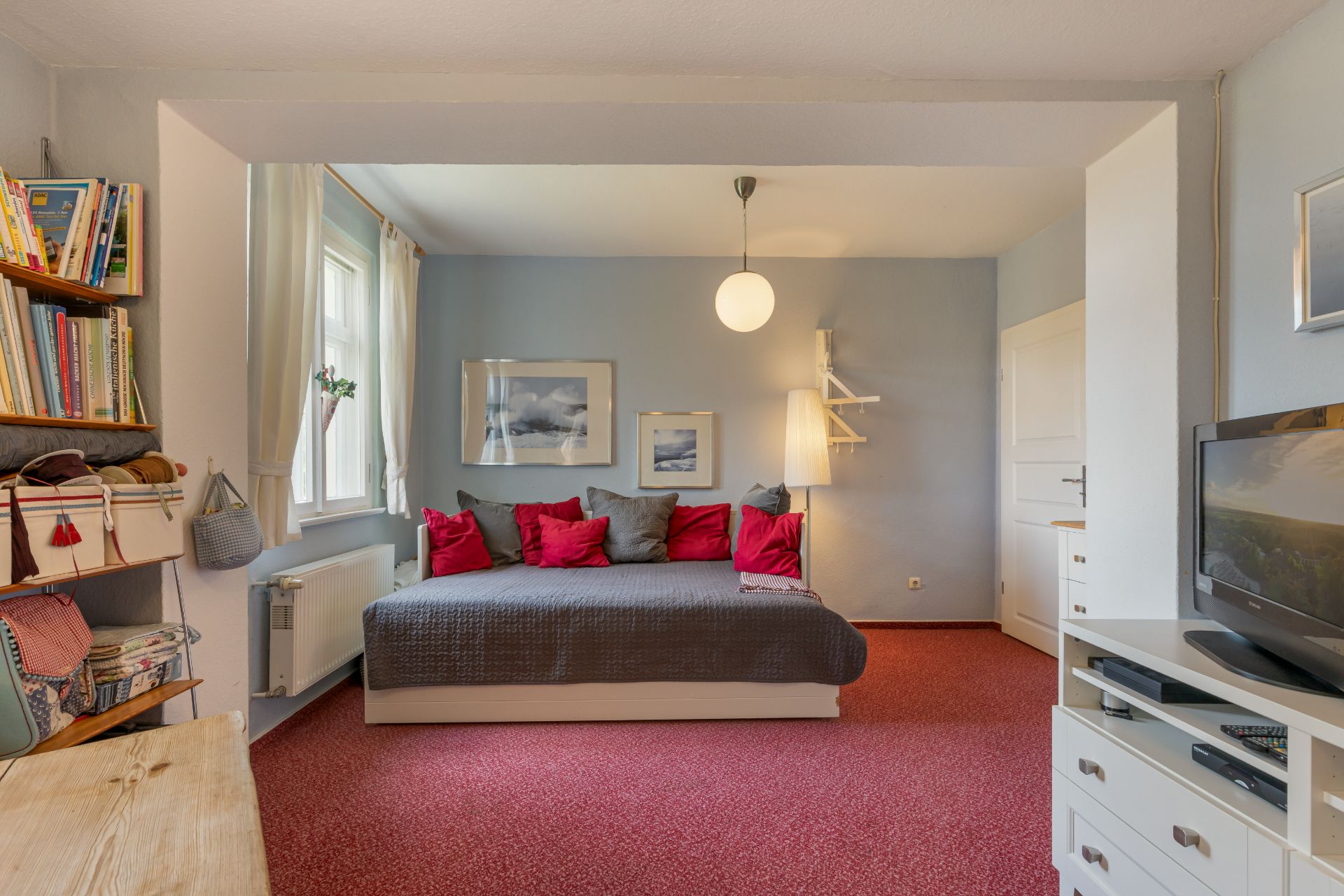 Wohnzimmer mit Bett, roten Kissen, Bücherregal, Fernseher und rotem Teppichboden.