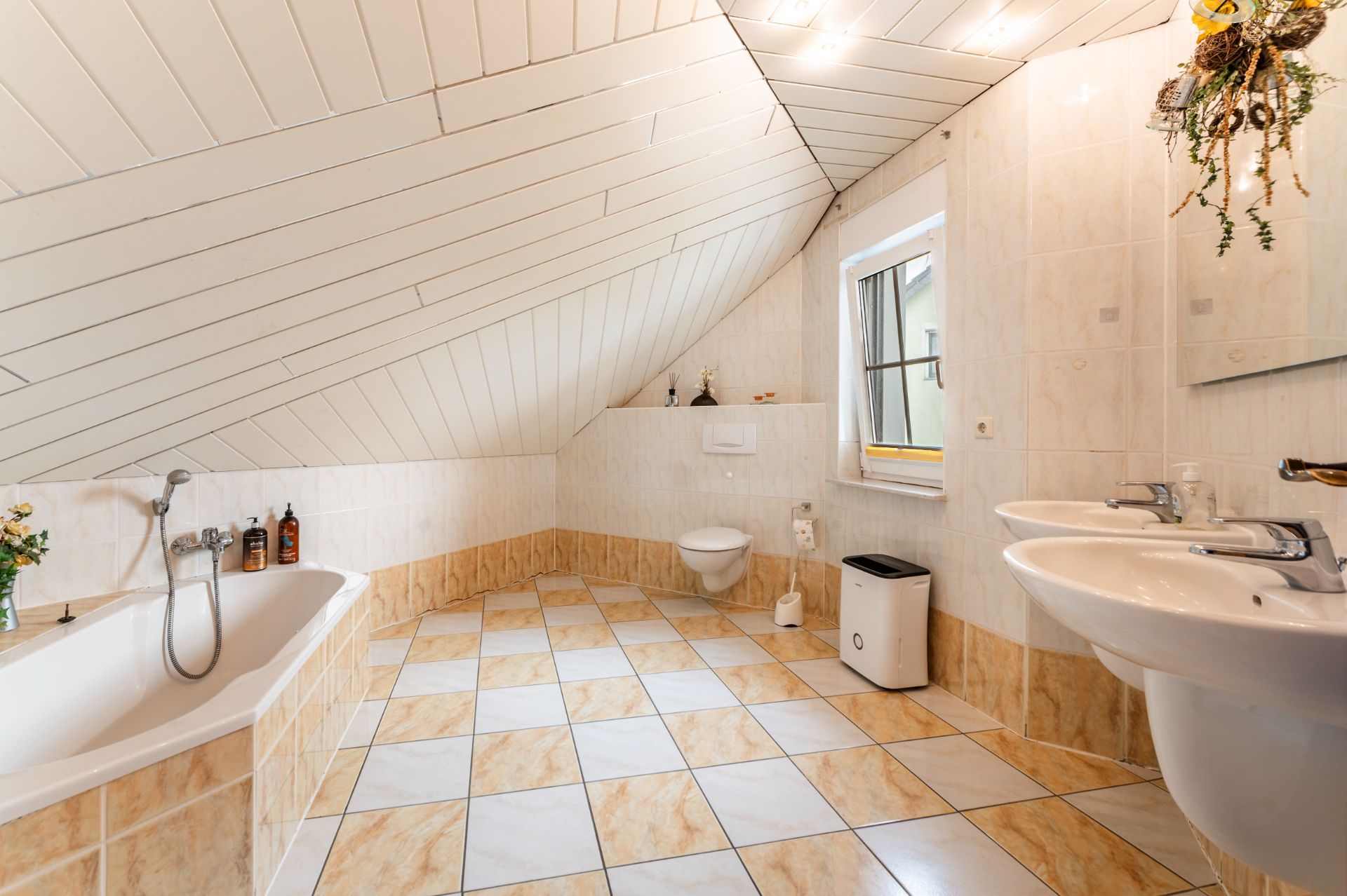 Helles Badezimmer mit Badewanne, zwei Waschbecken, WC und Fenster unter einer abgeschrägten Decke.