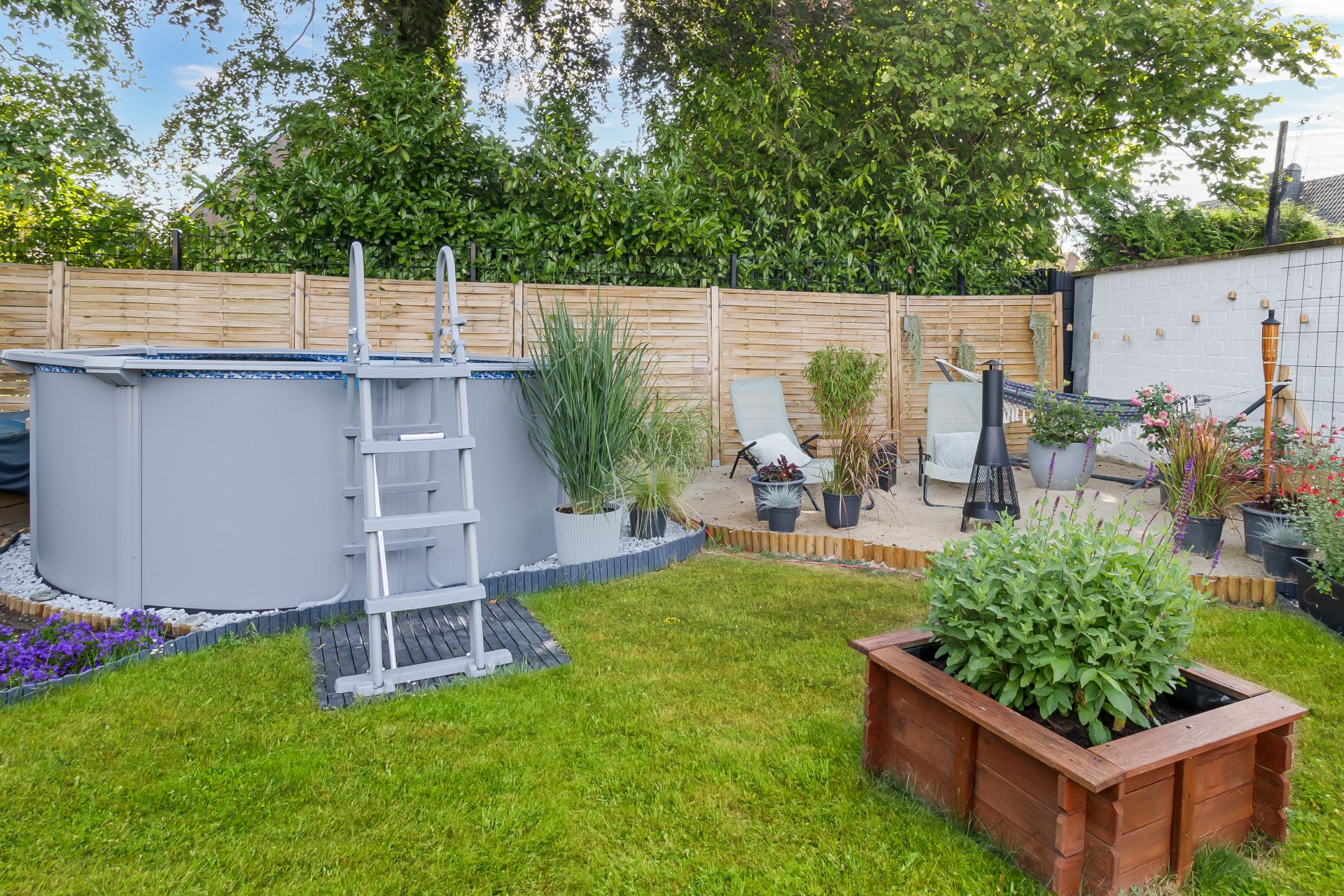 Garten mit Aufstellpool, Sitzbereich mit Stühlen und Kübelpflanzen auf Rasen.