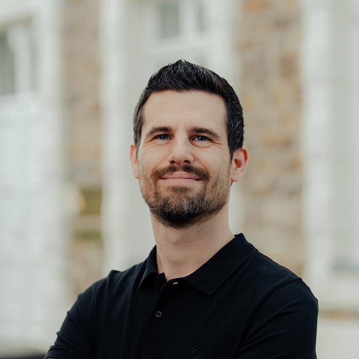 Ein Mann mit dunklem Bart und schwarzem Polo steht mit verschränkten Armen draußen.