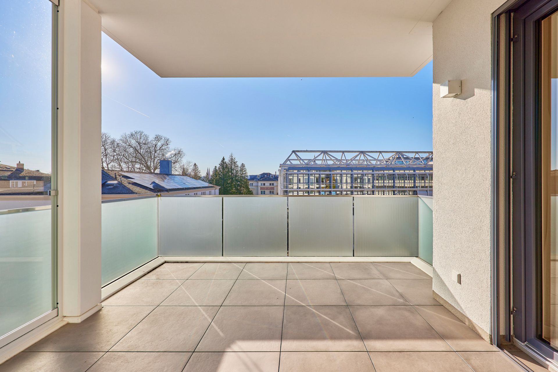 Großer, überdachter Balkon mit Glasbrüstung und Blick auf Gebäude und Bäume.
