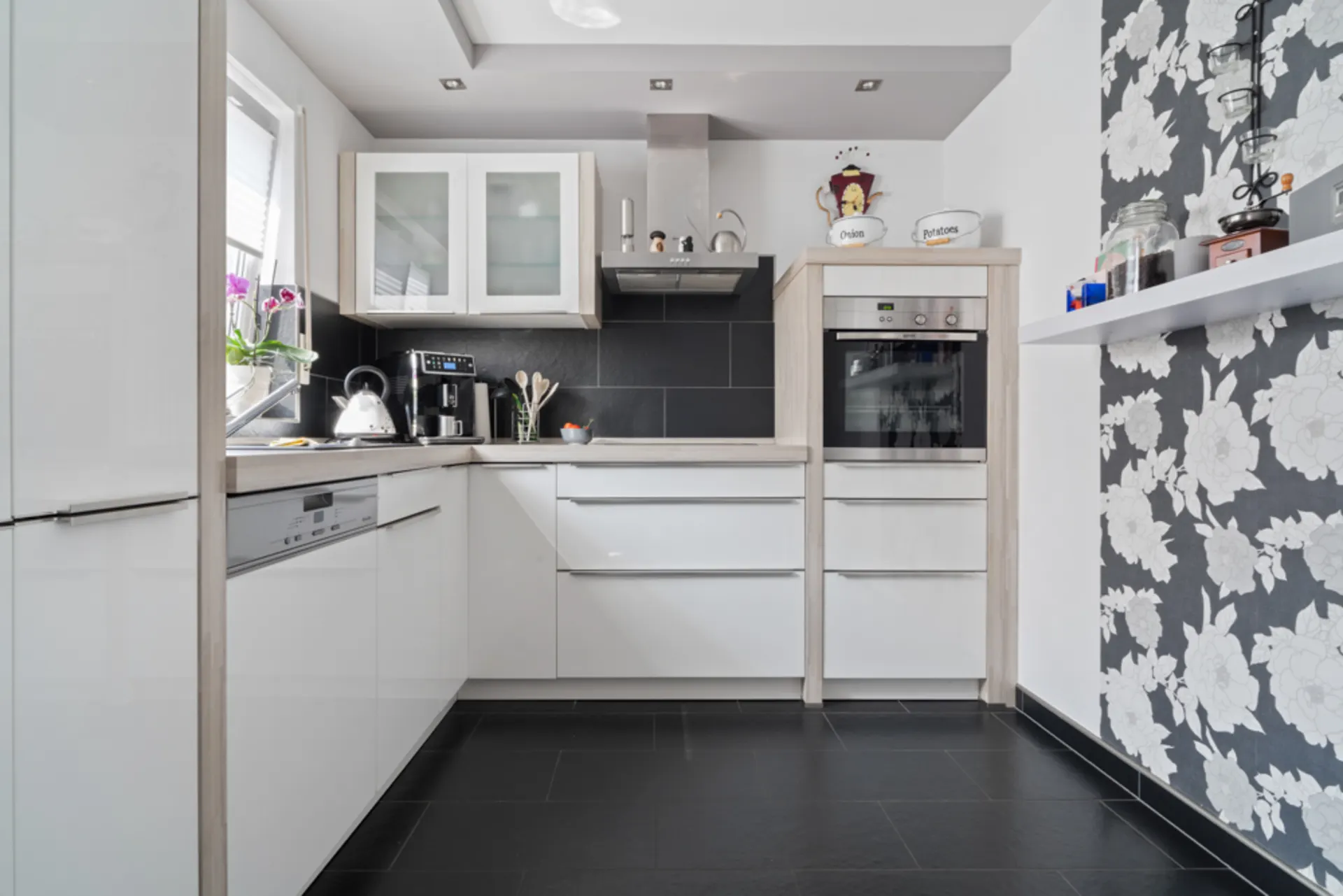Helle, moderne Küche mit weißen Schränken, schwarzen Fliesen und floral gemusterter Tapete.