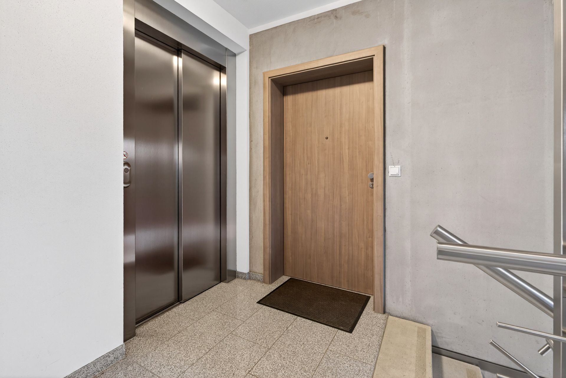 Hausflur mit Aufzug, Wohnungstür aus Holz und Treppengeländer.