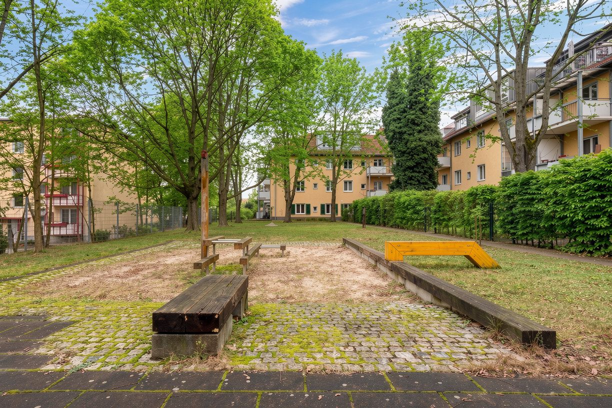Kleiner Spielplatz mit Holzbänken und Sandfläche zwischen Wohngebäuden und Bäumen.