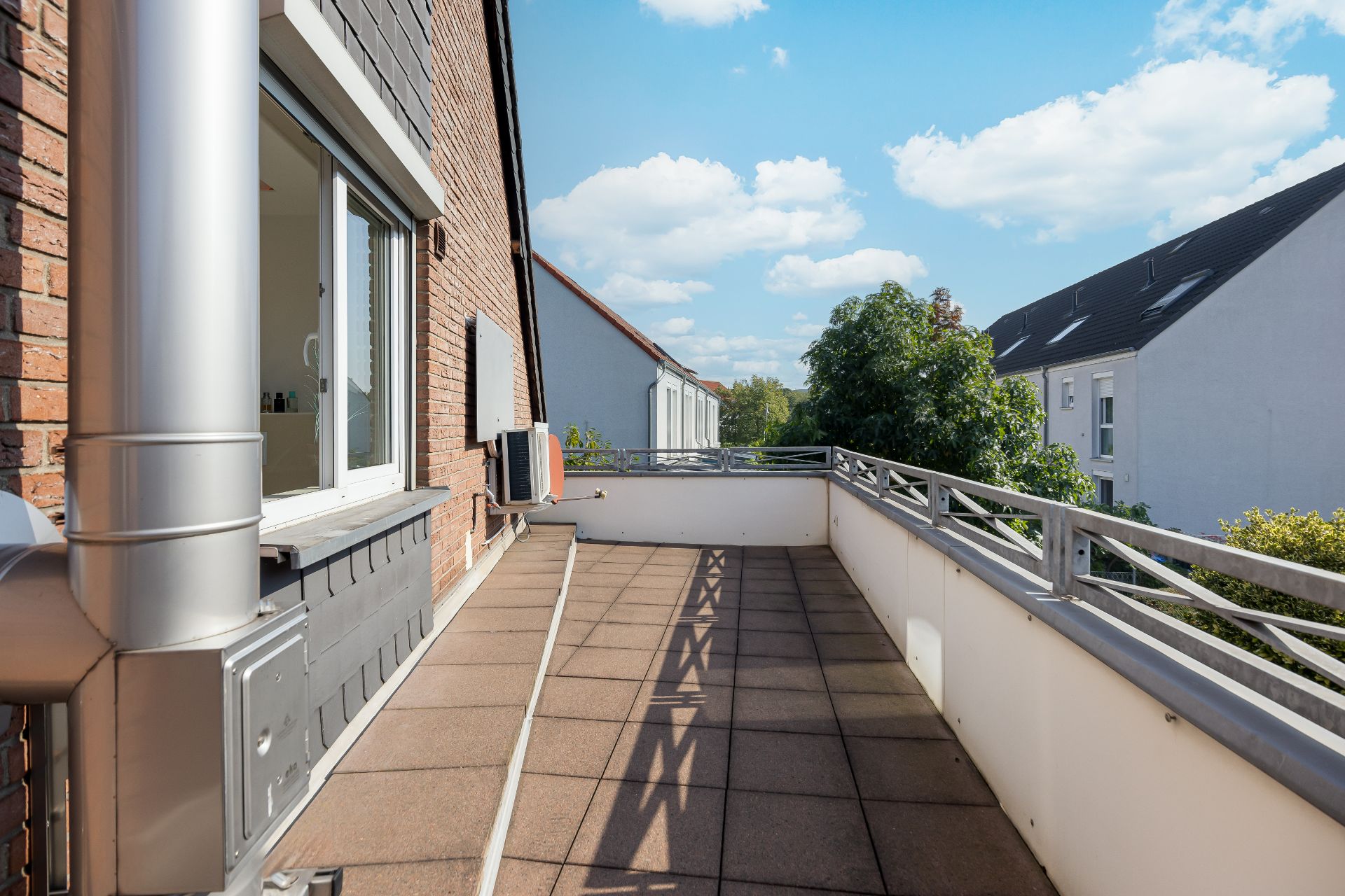 Großer Balkon mit Fliesenboden und Geländer, Blick auf Nachbarhäuser und Bäume bei sonnigem Wetter.