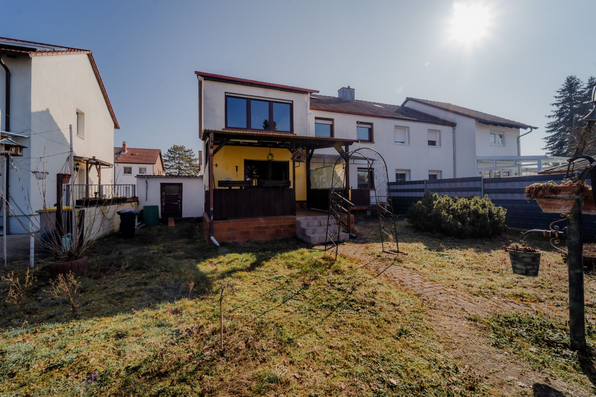 Rückseite eines Hauses mit Terrasse, Garten und angrenzenden Nachbarhäusern bei Sonnenschein.