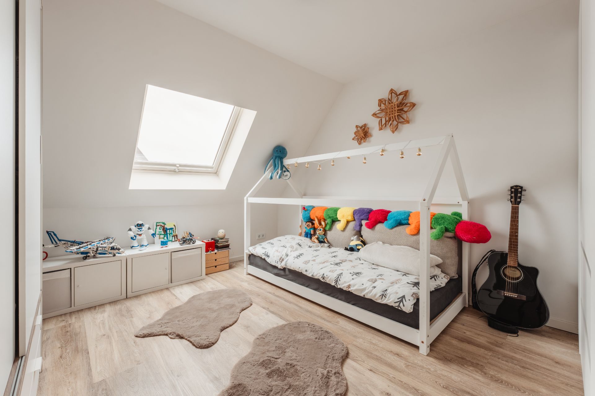 Helles Kinderzimmer mit Hausbett, vielen Kuscheltieren, Spielzeug, Gitarre und Teppichen auf Holzfußboden.