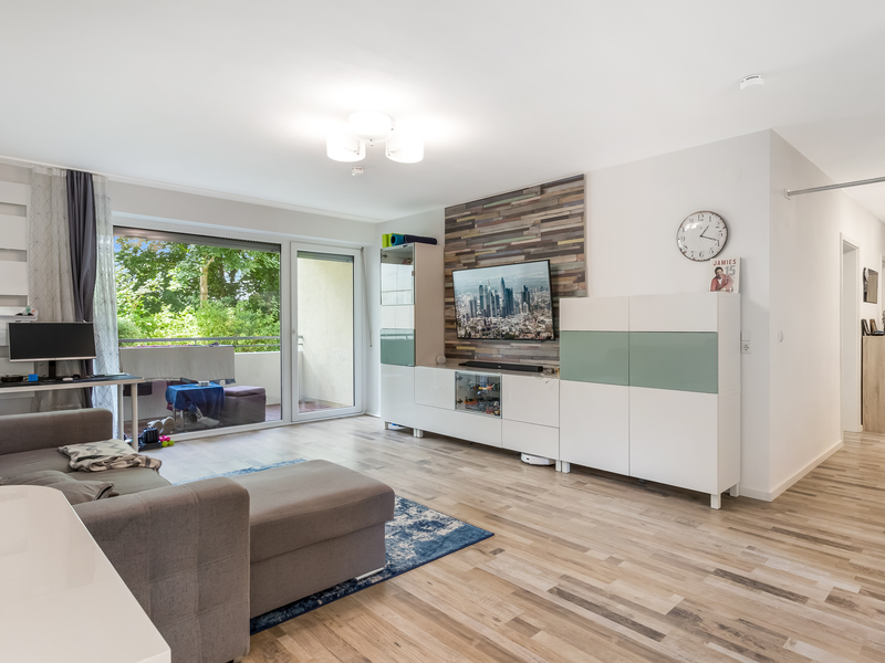 Modernes Wohnzimmer mit Sofa, Fernseher, Schrankwand und Zugang zu einem Balkon mit Blick ins Grüne.