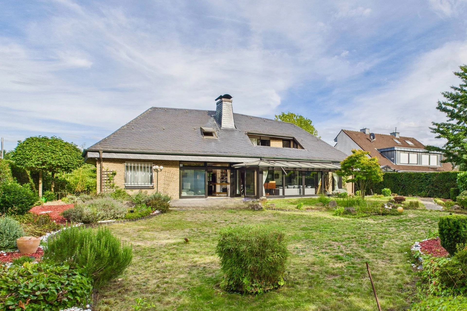 Zweistöckiges Haus mit großem Garten und Terrasse, umgeben von Sträuchern und Bäumen.