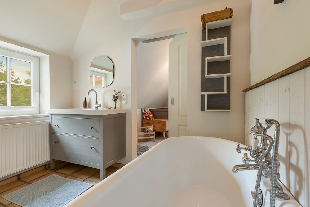 Helles Badezimmer mit freistehender Badewanne, Waschtisch, Fenster und Regal an der Wand.