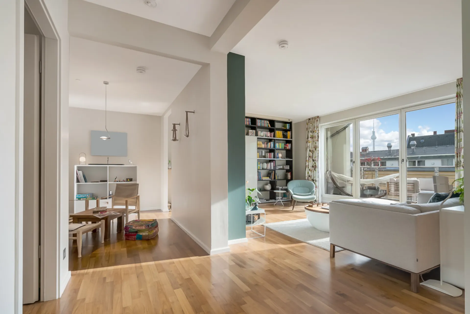 Helles Wohnzimmer mit Sofa, Sessel, Bücherregal und bodentiefen Fenstern zu einem Balkon.