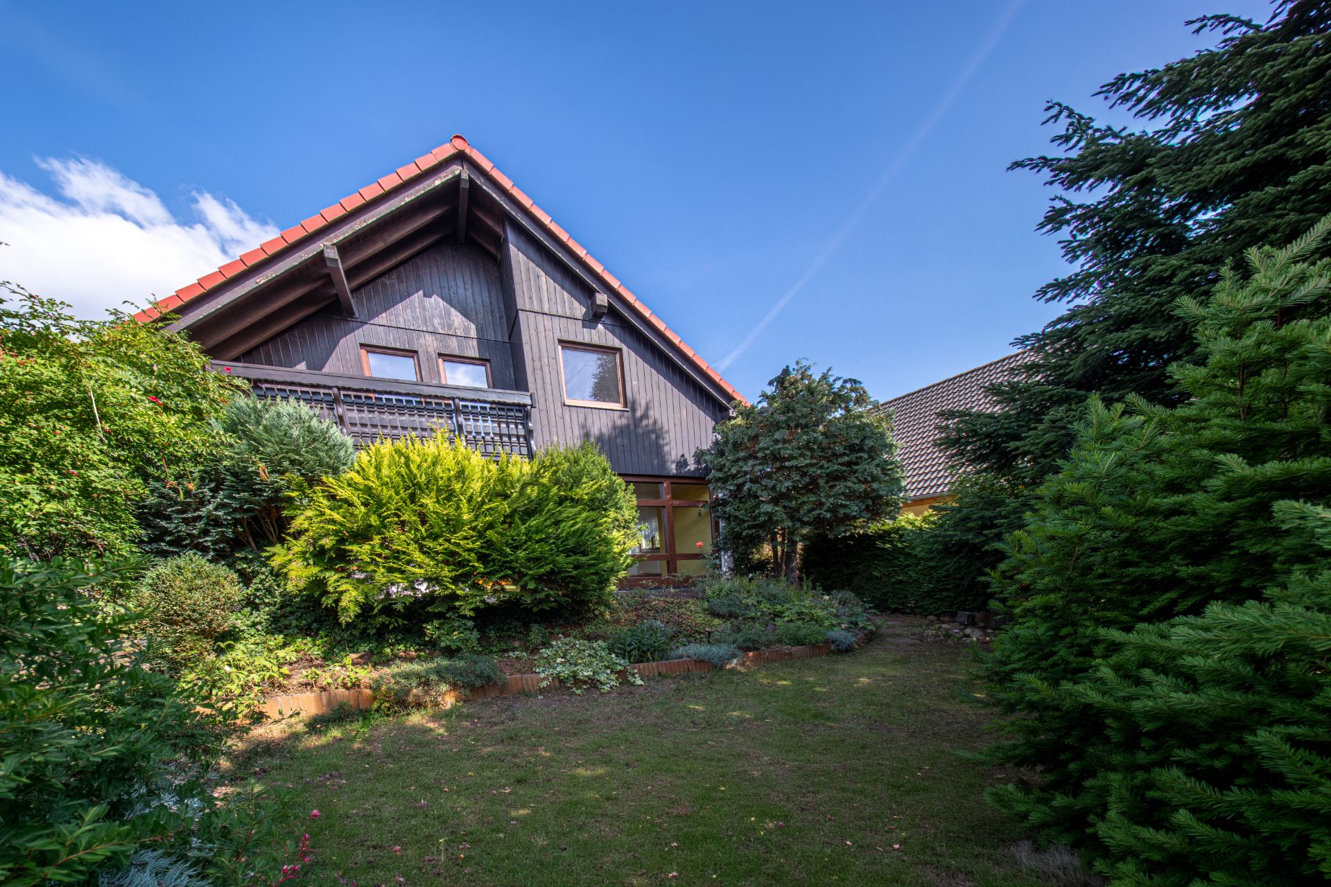 Haus mit dunkel verkleideter Fassade, großem Fenster und Garten mit verschiedenen Bäumen und Büschen.