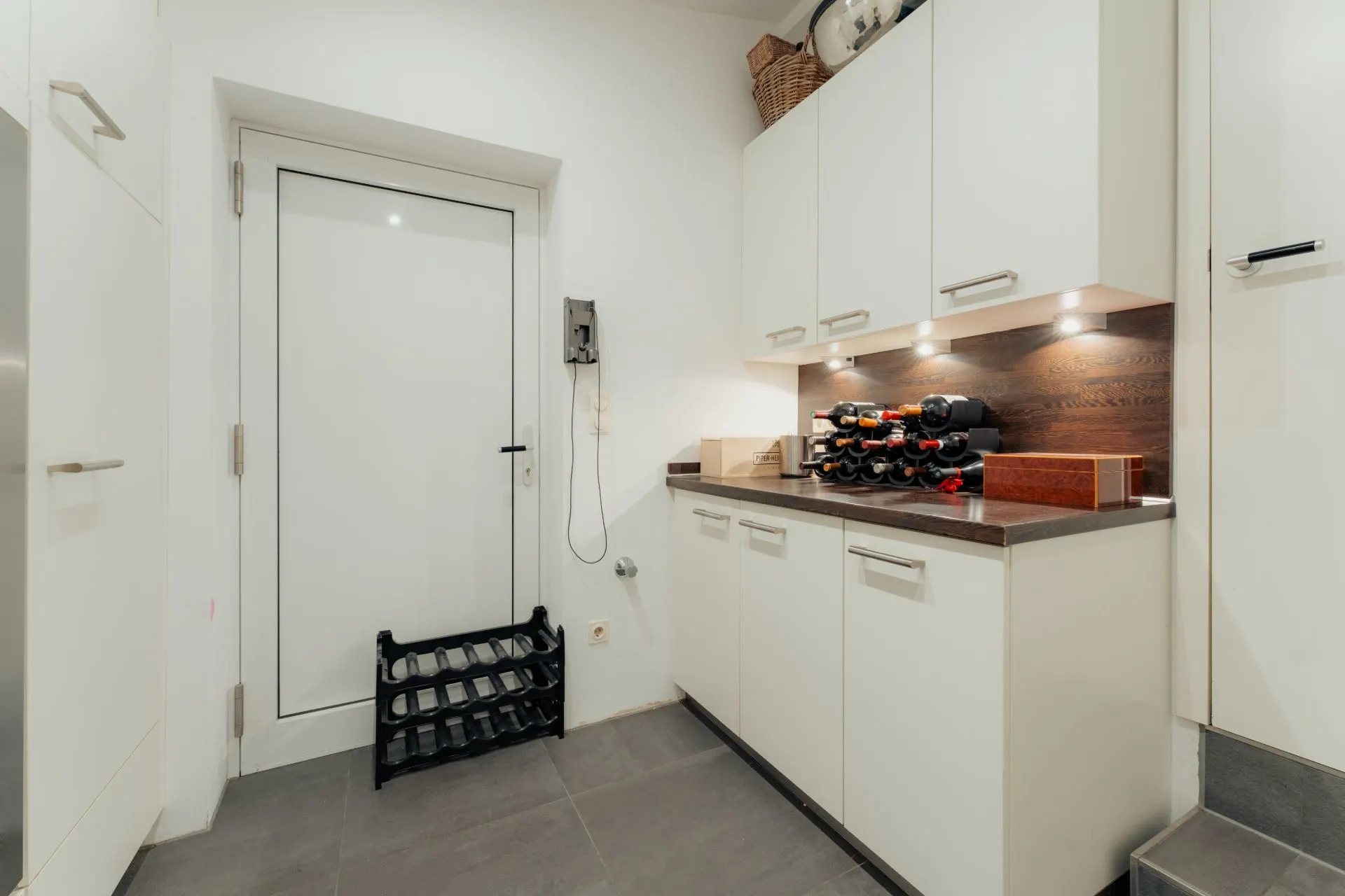 Moderner Hauswirtschaftsraum mit weißen Schränken, Weinregal und einer Tür.