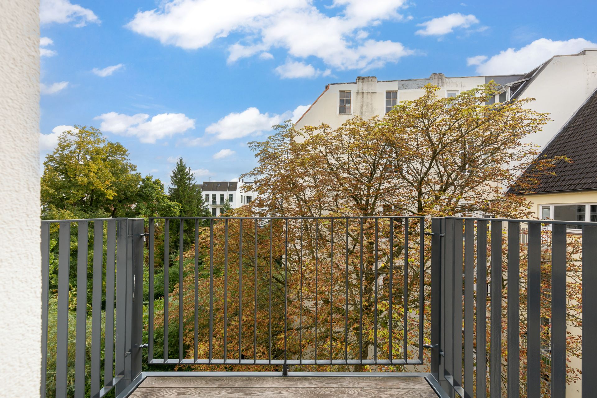 Balkon mit Metallgeländer, Blick auf Bäume und Nachbargebäude bei blauem Himmel.