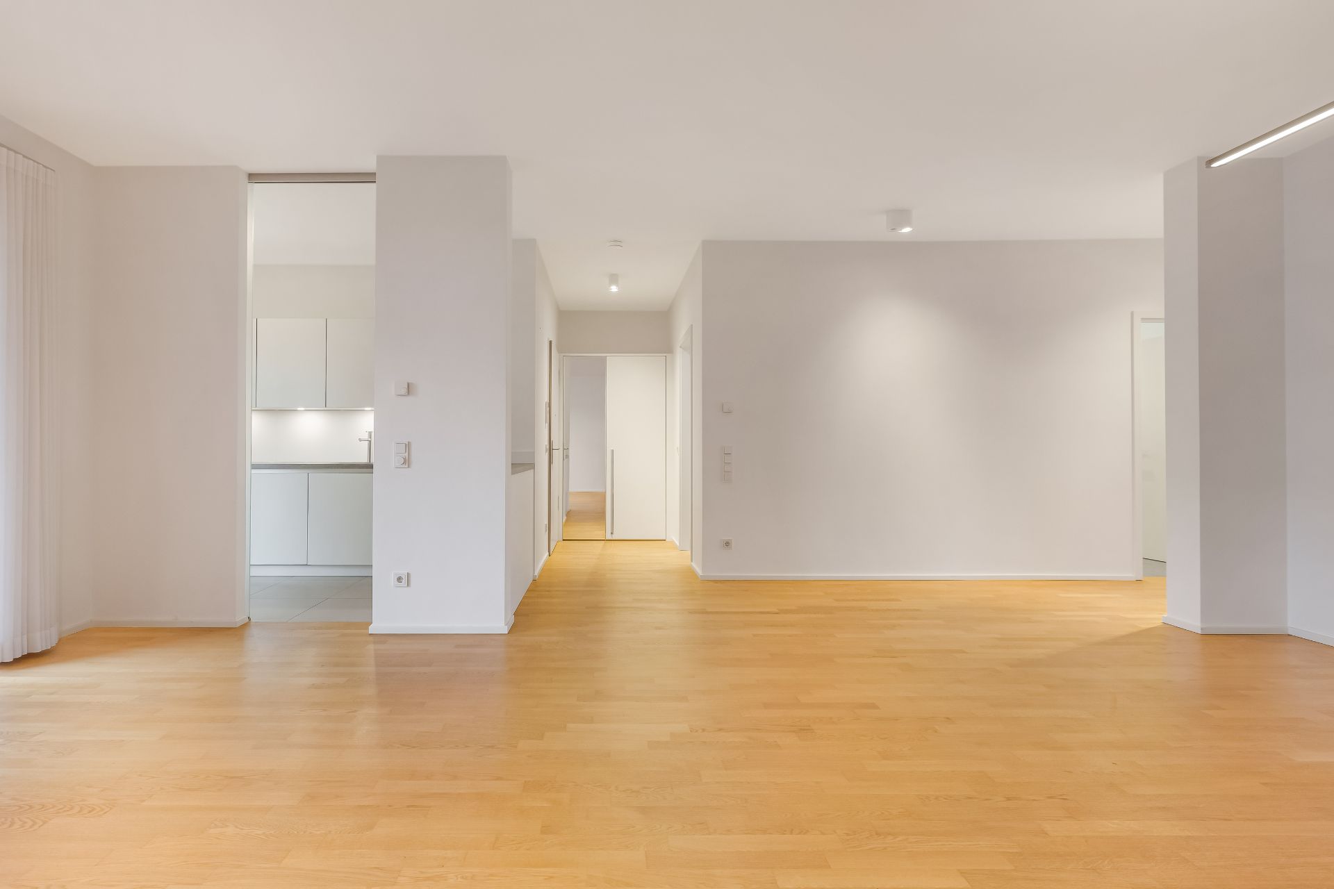 Heller, leerer Raum mit Holzfußboden und Blick in eine weiße Einbauküche.