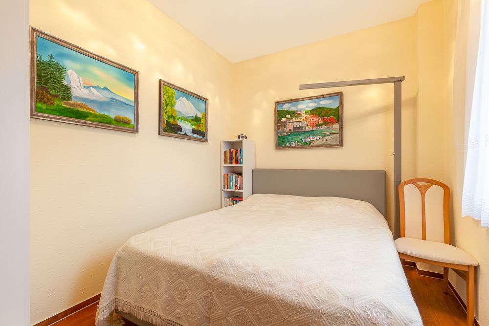 Kleines Schlafzimmer mit Doppelbett, Gemälden an der Wand, Bücherregal und Stuhl am Fenster.