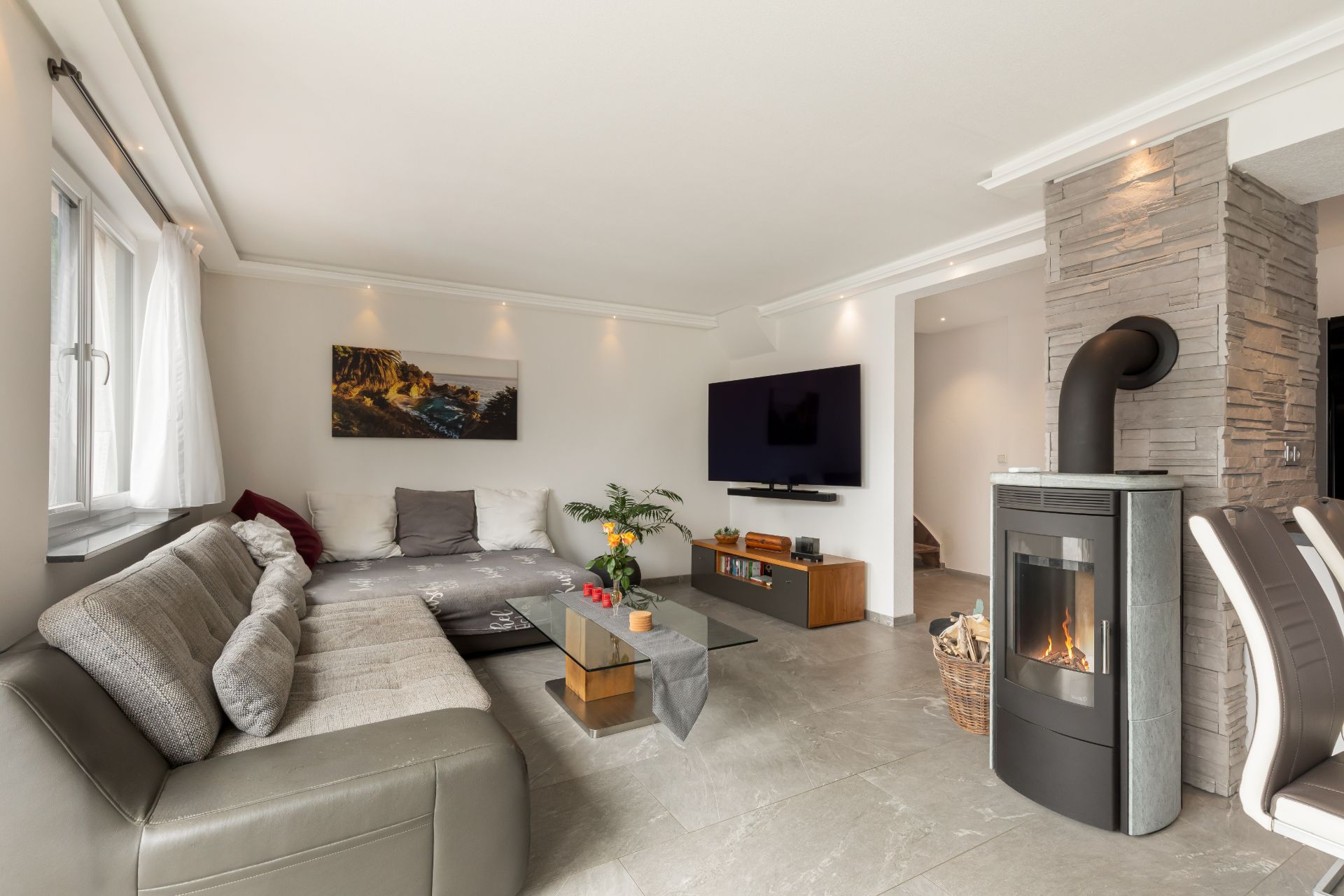 Modernes Wohnzimmer mit Ecksofa, Kaminofen, Fernseher und Couchtisch, daneben Fenster mit weißen Vorhängen.