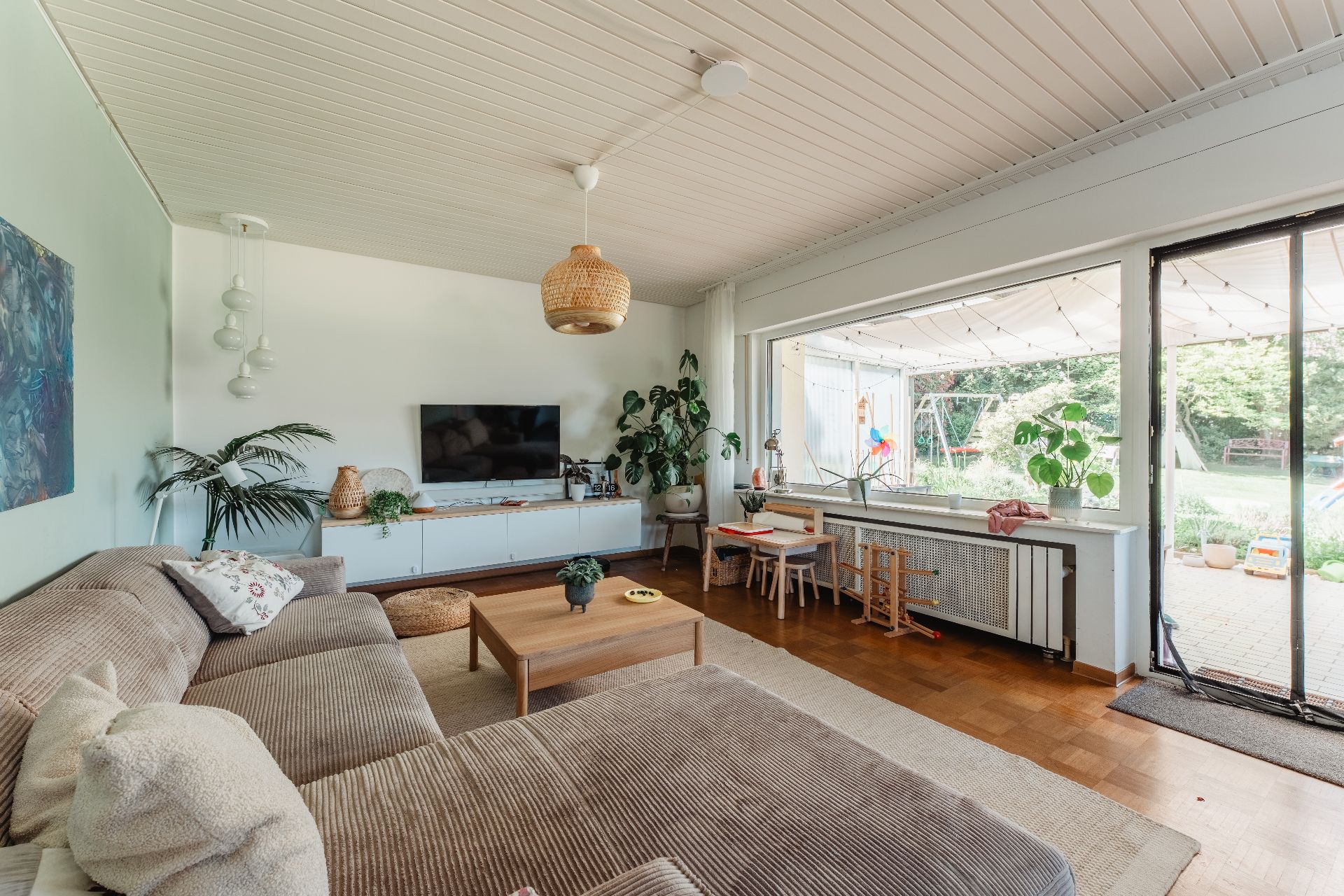 Wohnzimmer mit großem Sofa, Couchtisch, Fernseher, Pflanzen und Blick in den Garten durch große Fenster.