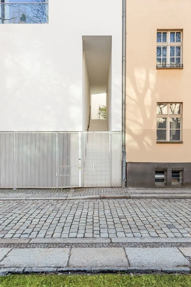 Moderne Hausfassade mit Durchgang und Treppe, daneben Fenster und gepflasterte Straße.