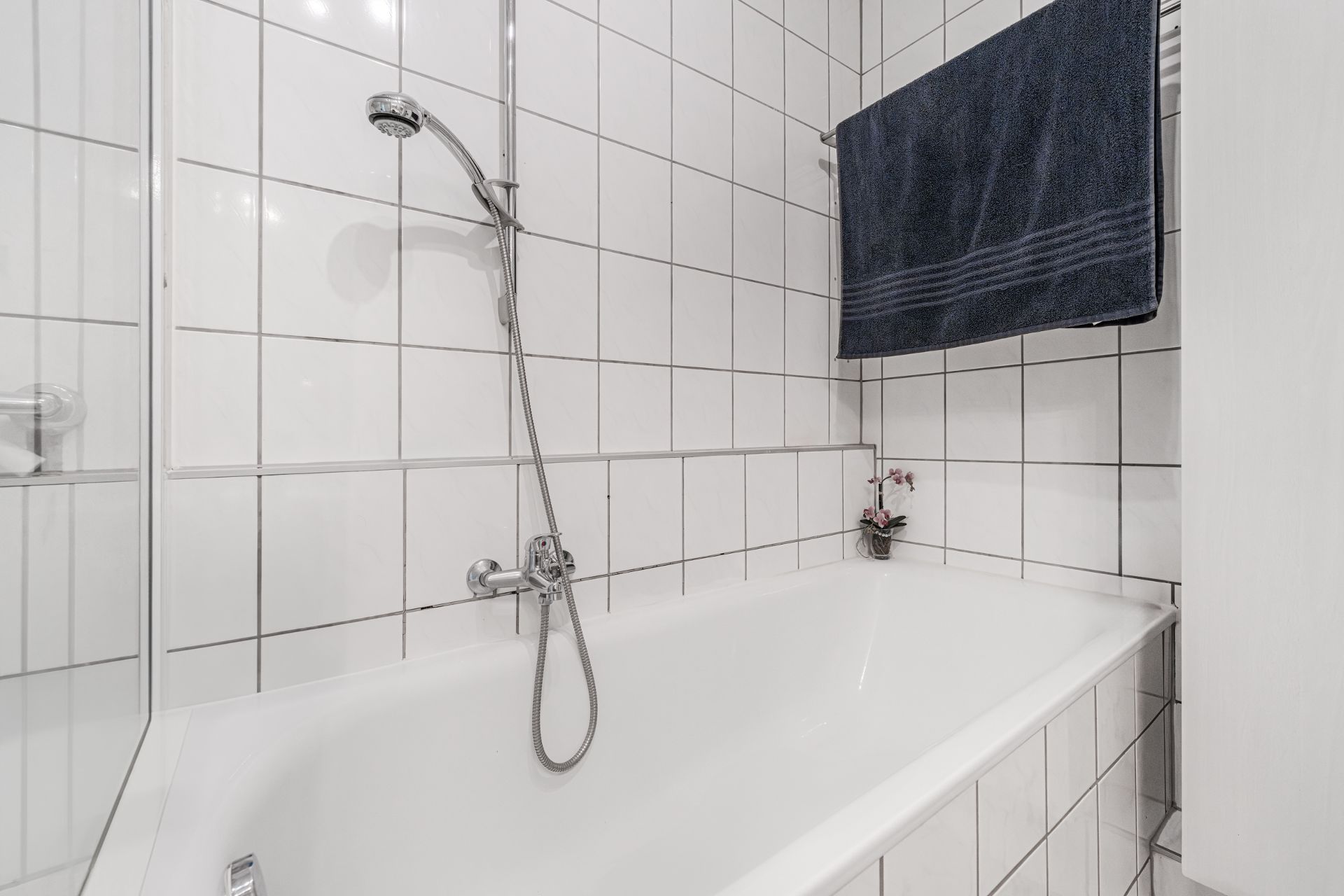 Weiße geflieste Badewanne mit Wanddusche, dunkelblauem Handtuch und kleiner Blume in der Ecke.