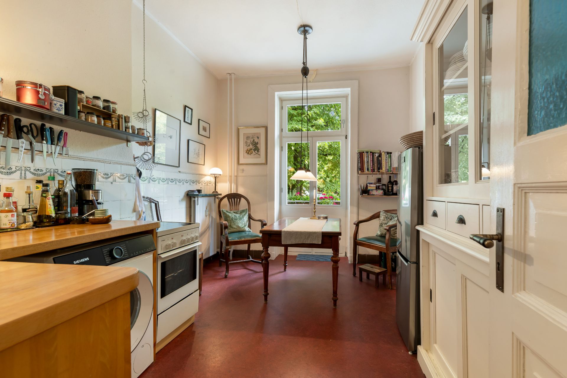 Helle Küche mit Esstisch, zwei Stühlen, Regal, Herd, Kühlschrank und Fenster mit Blick ins Grüne.