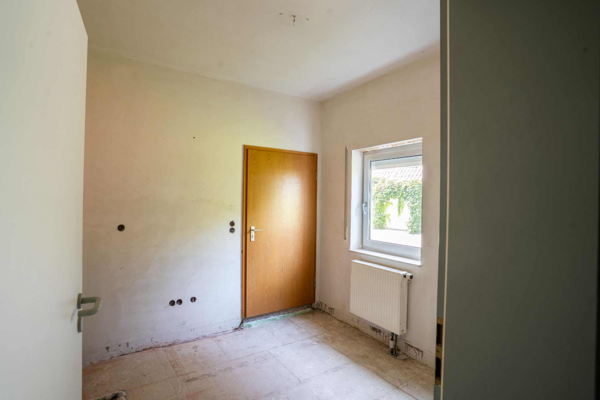 Leerer Raum mit Holztür, Fenster und Heizkörper, Boden ohne Belag.
