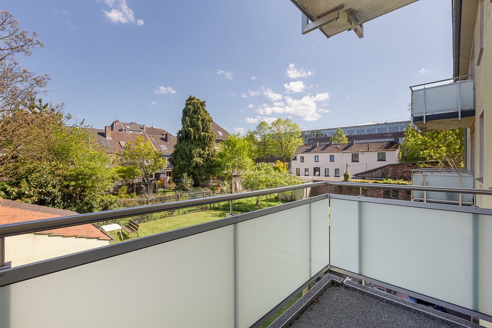 Balkon mit Glasgeländer, Blick auf Gärten und Wohnhäuser bei sonnigem Wetter.