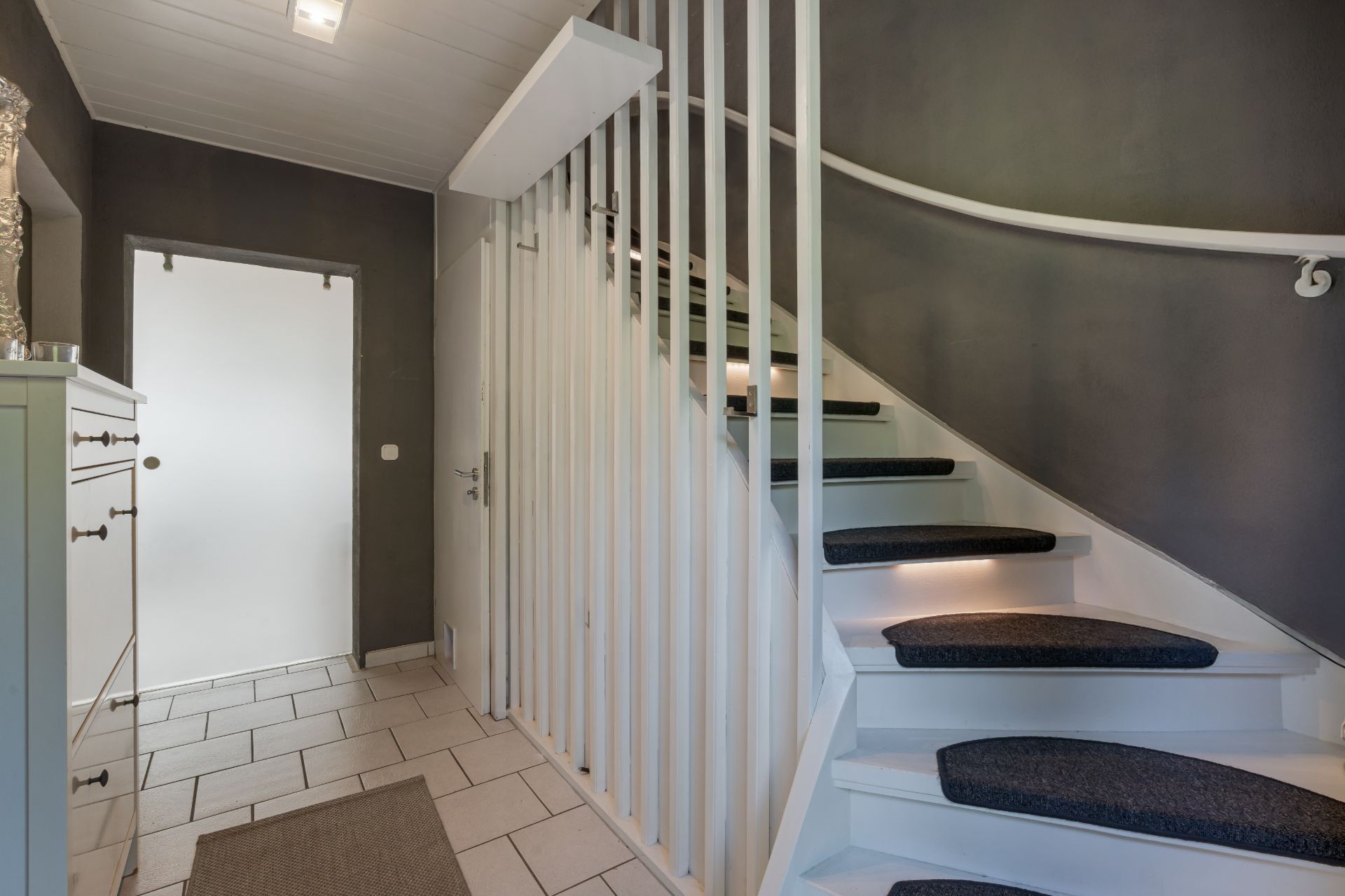 Heller Flur mit gefliesten Boden und weißer Treppe mit Stufenmatten.