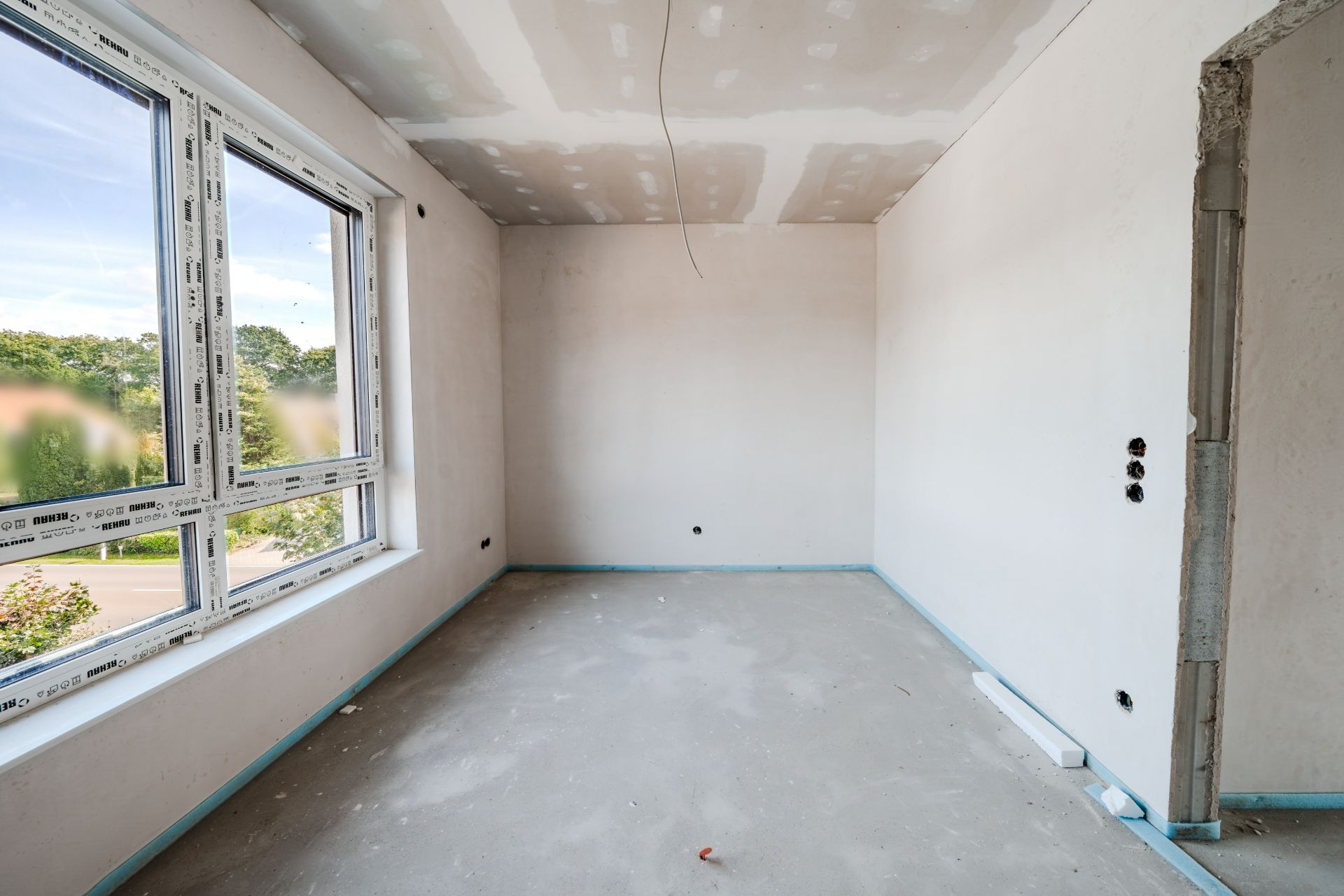 Helles, unfertiges Zimmer mit großen Fenstern und weißem Putz an Wänden und Decke.