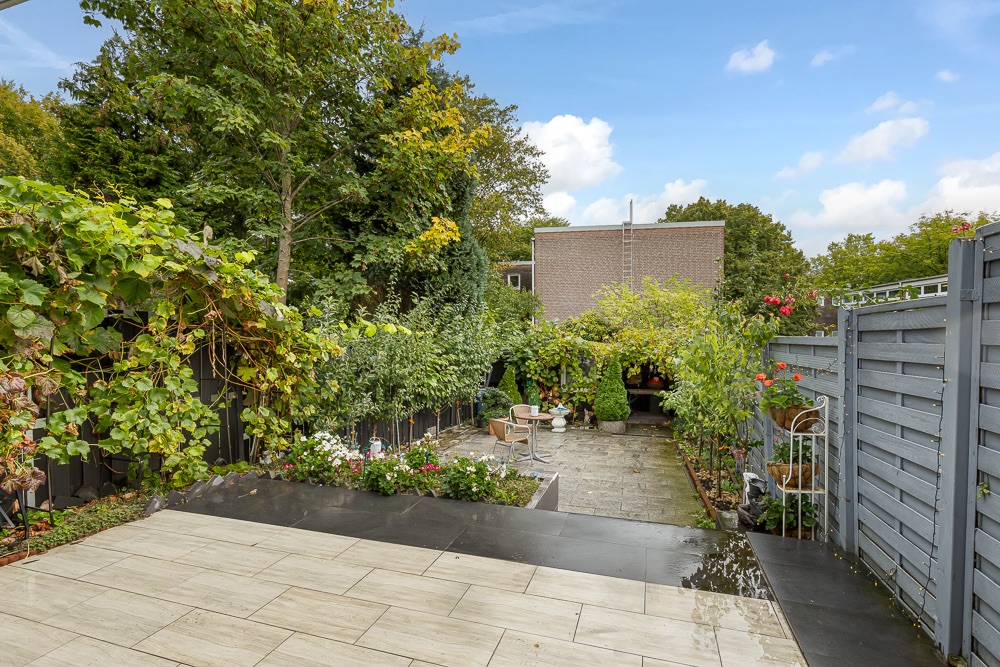 Gepflasterte Terrasse mit Garten, Bäumen und grauem Holzzaun, viele grüne Pflanzen sichtbar.