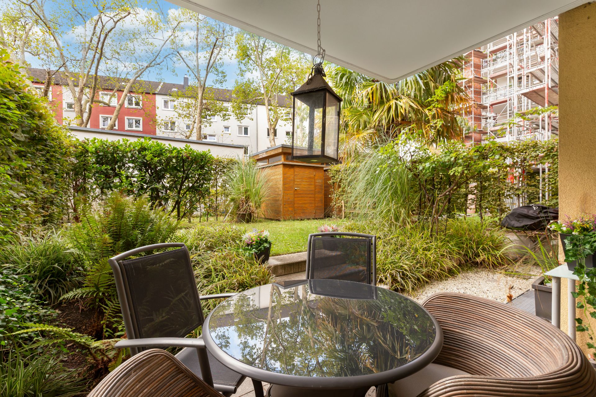 Terrasse mit rundem Glastisch, vier Stühlen und Blick auf Garten mit Gartenhaus.
