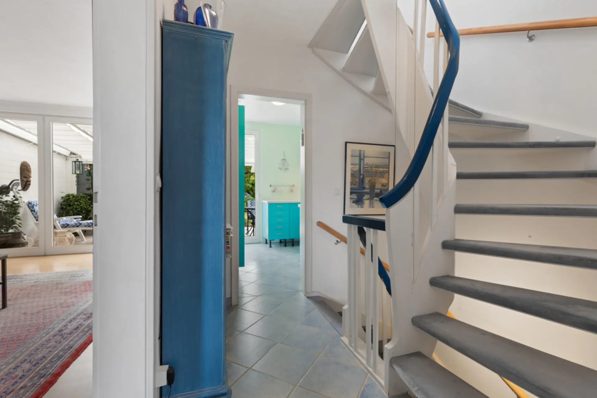 Flur mit blauer Treppe, blauem Schrank und Blick in ein helles Zimmer mit Fliesenboden.