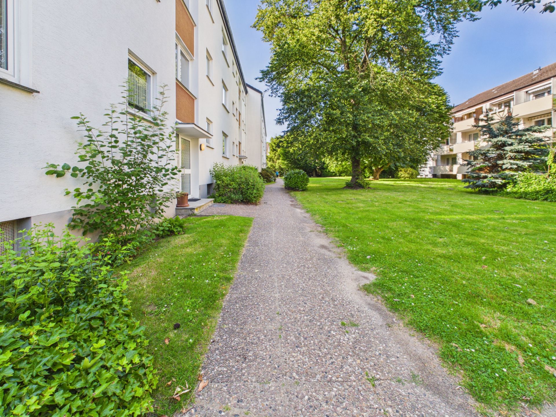 Gepflegter Gehweg neben einem Mehrfamilienhaus und großer grüner Wiese mit Bäumen.