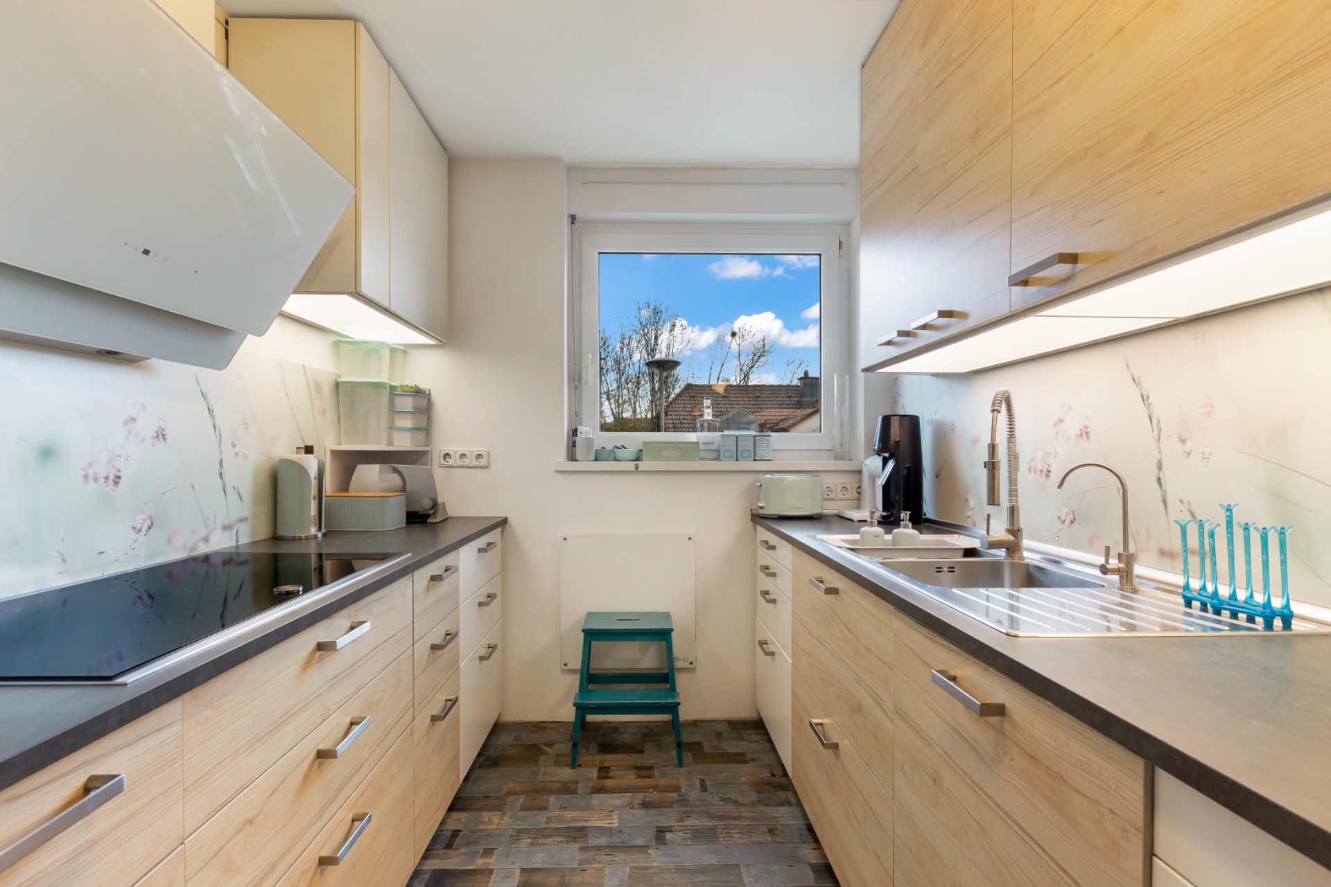 Moderne Küche mit hellen Holzmöbeln, Geräten, Fenster und Hocker auf gefliestem Boden.