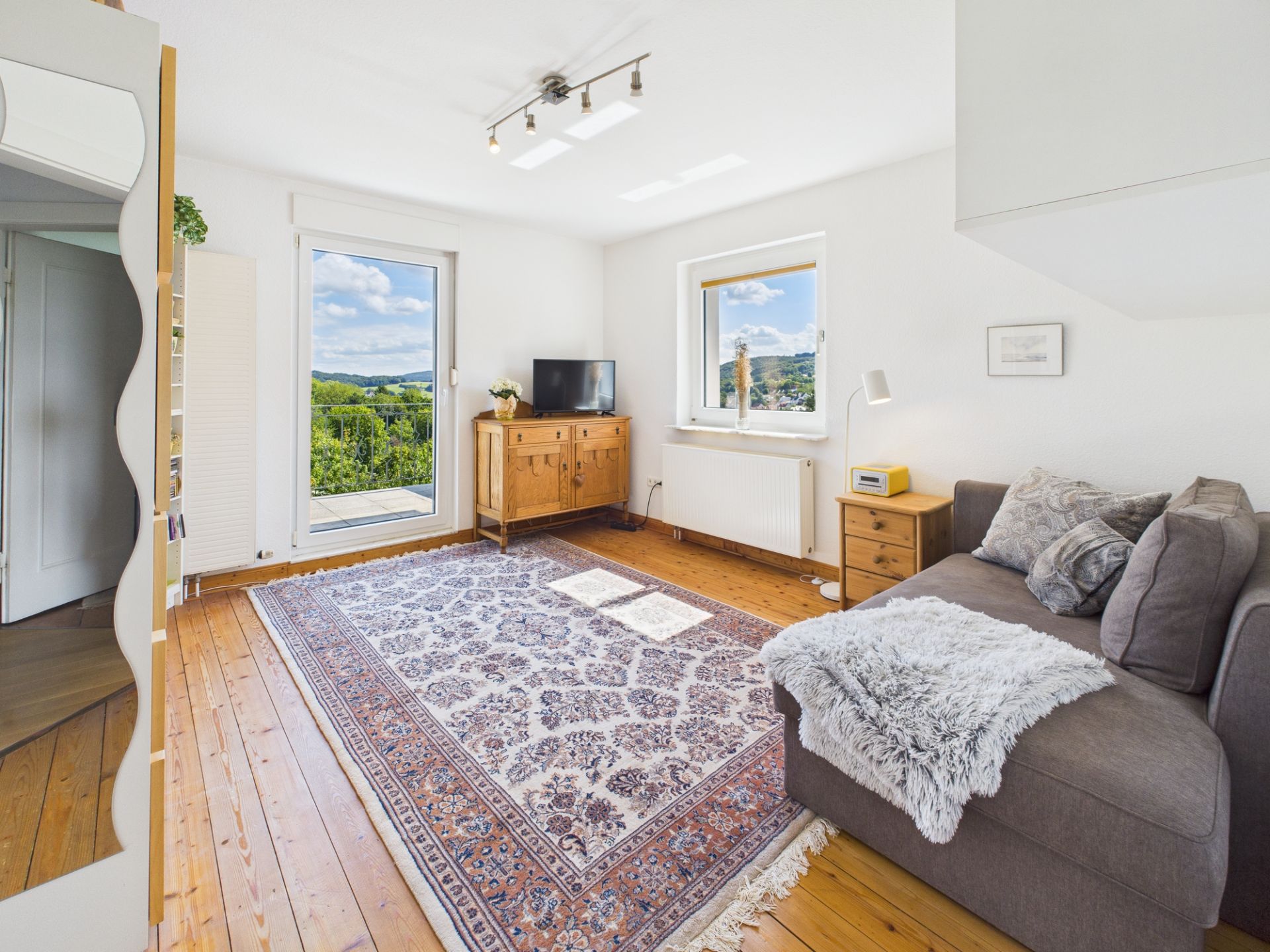 Helles Wohnzimmer mit großem Teppich, Sofa, Kommode, TV und Blick durch große Fenster ins Grüne.