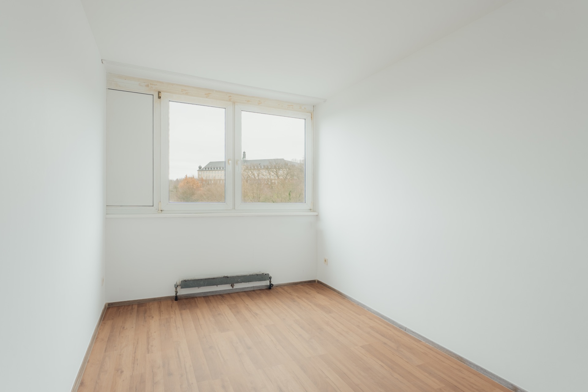 Leerer, heller Raum mit weiß gestrichenen Wänden, Holzfußboden und großem Fenster.