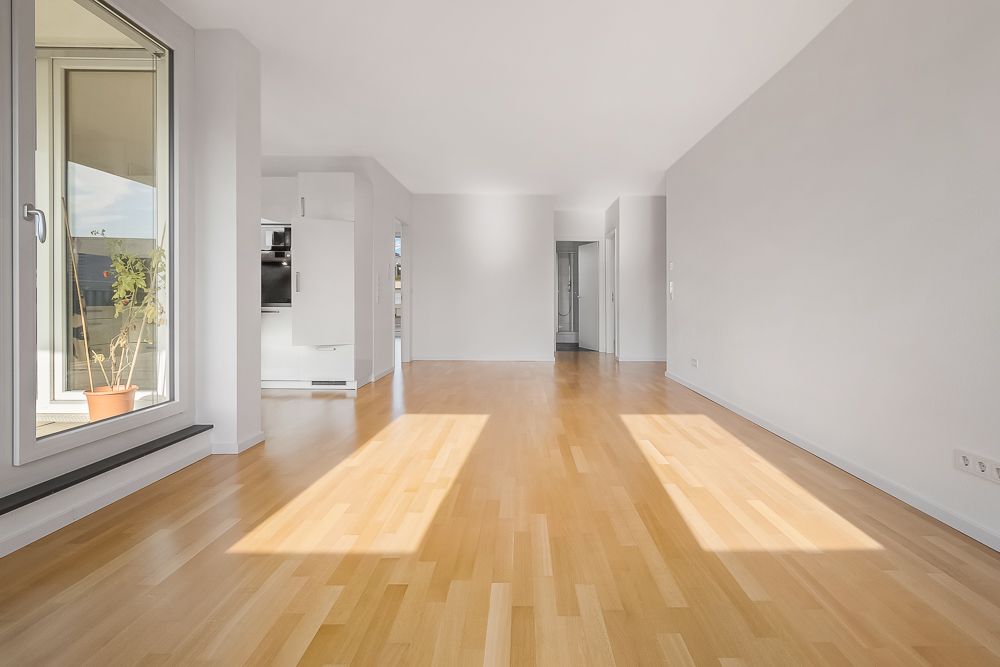 Helles, leeres Wohnzimmer mit Holzfußboden, großen Fenstern und Blick zur offenen Küche.