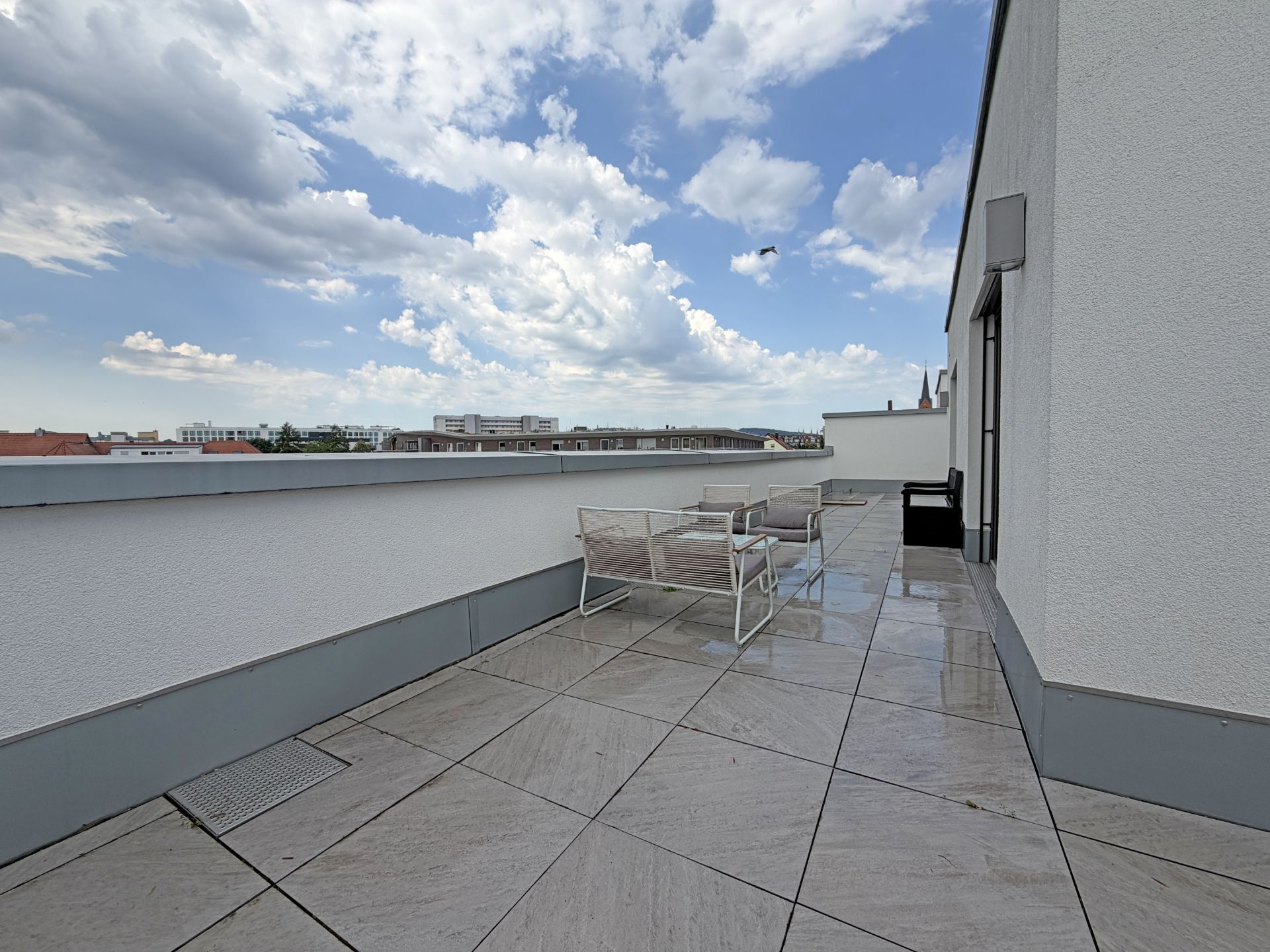 Große Dachterrasse mit Sitzmöbeln und Fliesenboden, Blick auf die Stadt und bewölkten Himmel.