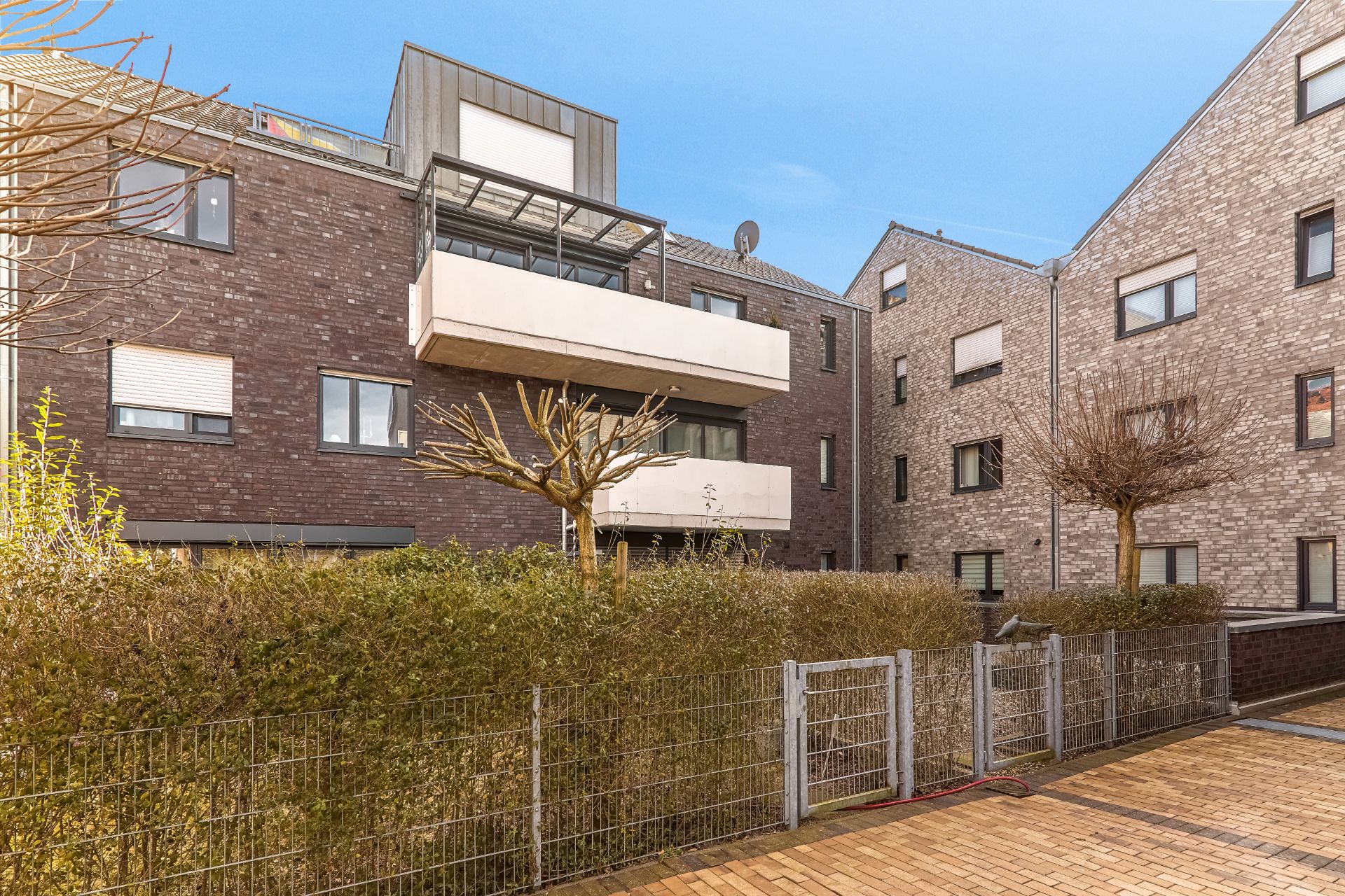 Mehrfamilienhaus mit mehreren Balkonen, Bäumen und einem Zaun im Vordergrund.