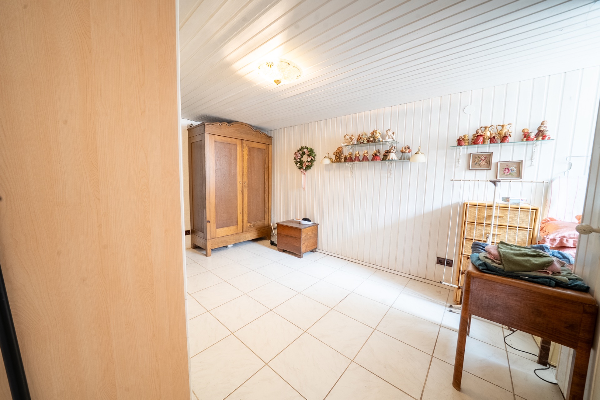 Helles Zimmer mit Fliesenboden, Holzschrank, Kommode und Wandregalen mit Dekoration.