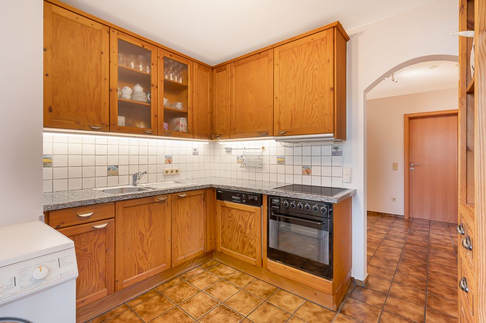 Küche mit Holzschränken, Herd, Spülmaschine und Fliesenboden, daneben Flur mit Holztür.