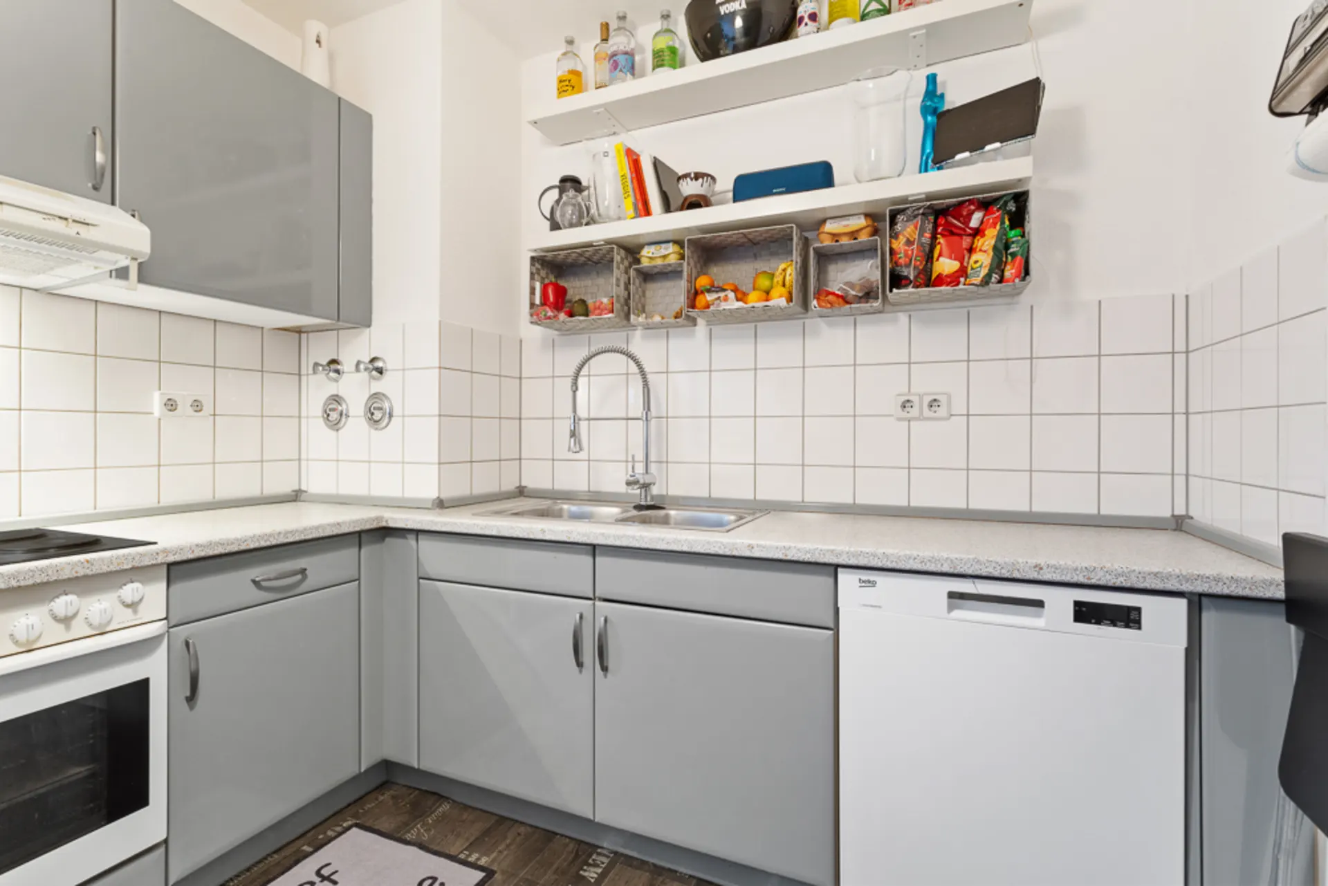 Helle, moderne Küche mit grauen Schränken, Spüle, Herd, Geschirrspüler und Wandregalen mit Lebensmitteln.