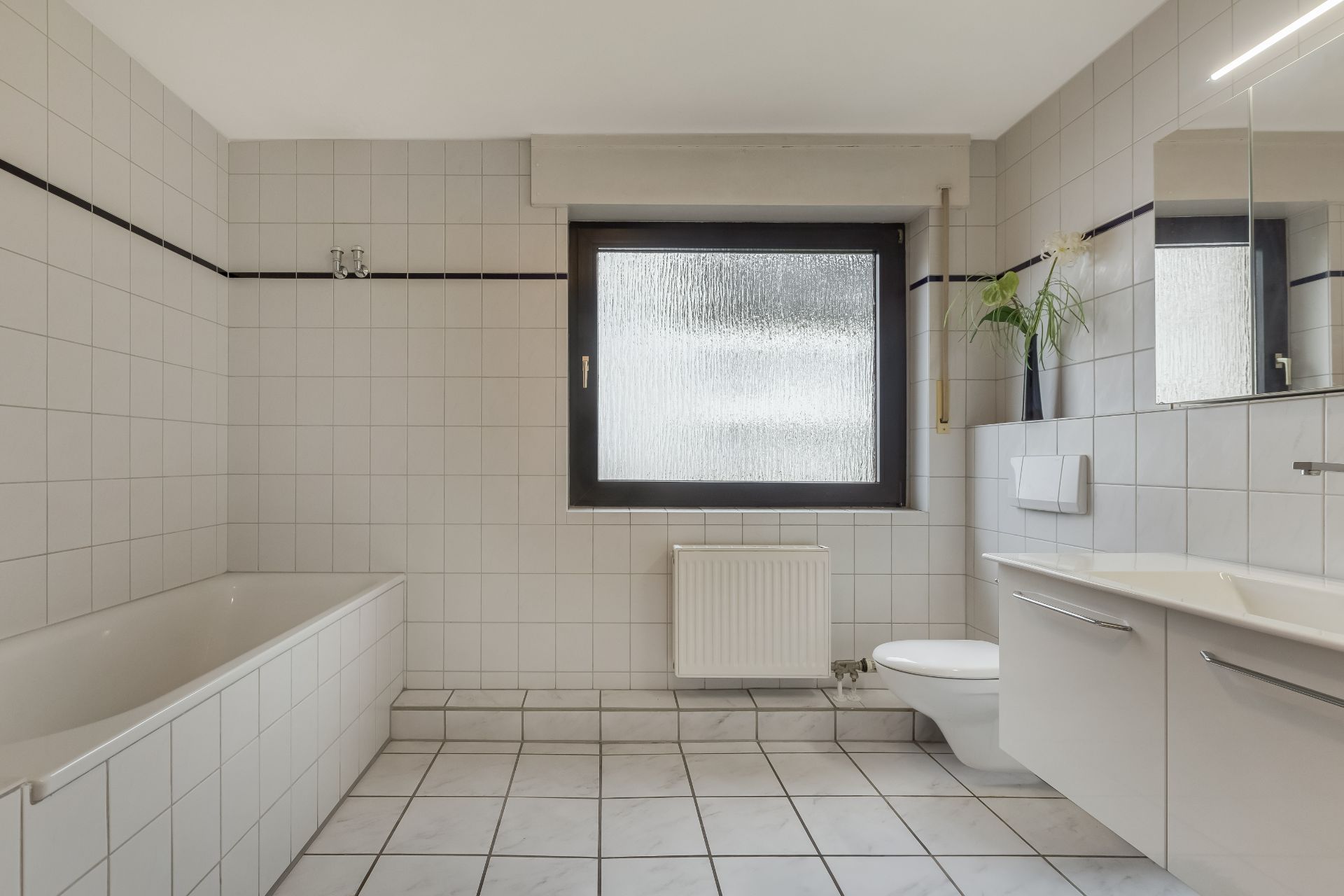 Helles Badezimmer mit Badewanne, Toilette, Waschbecken, Fenster und weiß gefliesten Wänden.